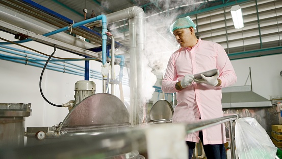 Asian engineers or technicians wearing hairnet and sterile clothing, inspect the working system and temperature large juice boiler via tablet in production line at bottled fruit juice processing plant. Concept of Industrial, manufacturing, factory.