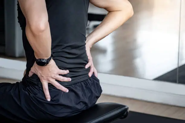 Photo of Sport man feeling lower back pain or spine pain while sitting on workout bench in fitness gym. Male athlete suffering from sport injuries symptoms.