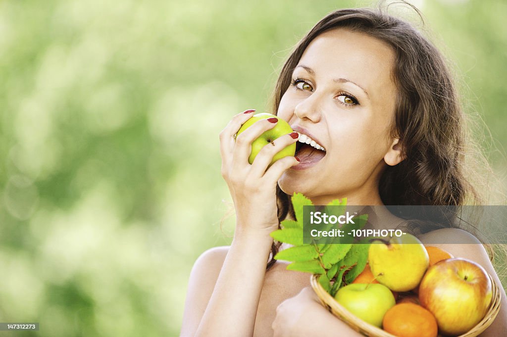 portrait of young attractive brunette woman eating apple Portrait of young attractive dark-haired smiling woman holding basket full of fruits and eating apple at summer green park. Apple - Fruit Stock Photo