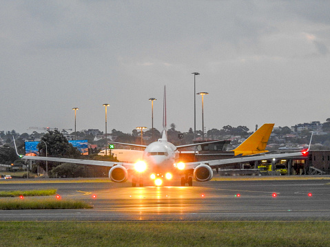 A Qantas Boeing B737-838 plane, VH-VXS, taxiing after having just landed from Melbourne on flight QF474 at Sydney Kingsford-Smith Airport.  In the distance is a DHL - Atlas Air Boeing B747-46N, registration N453PA, having been towed to the parking area near the fire station.  Two parked fire engines are visible on the right.  The B747 arrived on 9 March 2023 and as at 13 March 2023, has not departed.  This image was taken from near Shep's Mound, a public viewing area off Ross Smith Avenue, on an overcast evening on 12 March 2023.