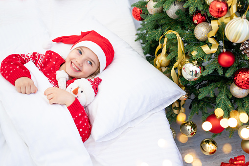 a child girl woke up under a Christmas tree in a red sweater and Santa Claus hat on New Year's Eve or Christmas in a white bed with a snowman in her arms