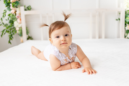funny baby girl six months old lies in a bright beautiful room on a white bed in a lace bodysuit