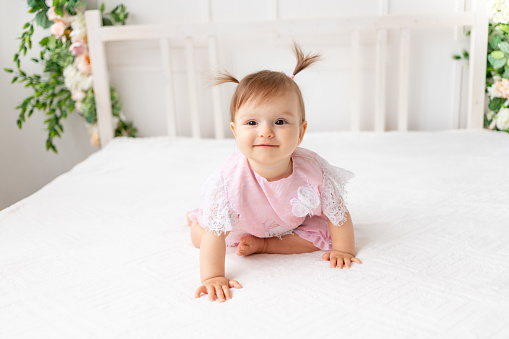 funny baby girl six months old lies in a bright beautiful room on a white bed in a lace bodysuit and smiles
