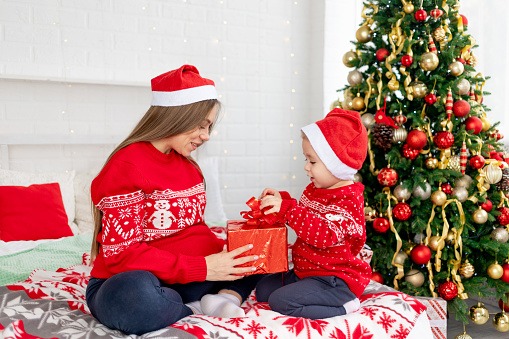 a pregnant woman with a baby boy in a red sweater and hats give gifts under the Christmas tree at home on the bed and rejoice in the new year and Christmas