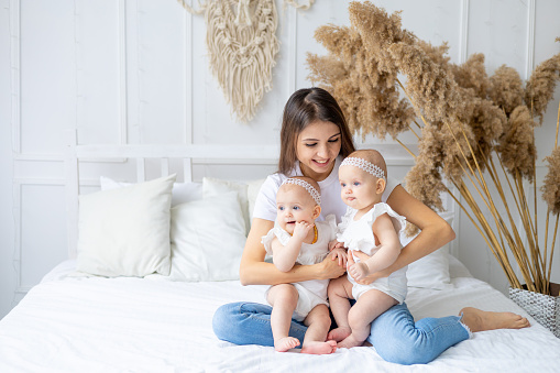 happy young mom with twin girls on the bed at home in cotton clothes in a bright bedroom, mom's love and care for small children
