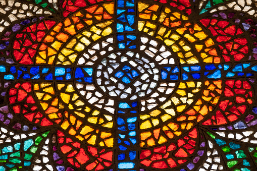 A dim old church interior lit by suns rays penetrating through a colorful stained glass window in the pattern of a crucifix reflecting colours on the floor and a speech pulpit