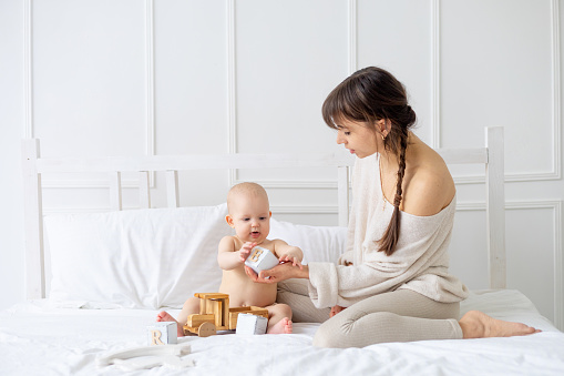 a young mother plays with a baby in diapers with wooden toys on a bright bed at home and smiles