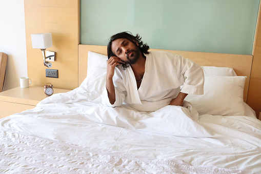 Stock photo showing close-up view of sat up man wearing towelling bathrobe in double bed whilst leaning head on hand.