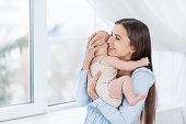mom holds a newborn baby girl in a cotton suit at the window of the house hugging and kissing him, happy motherhood or family