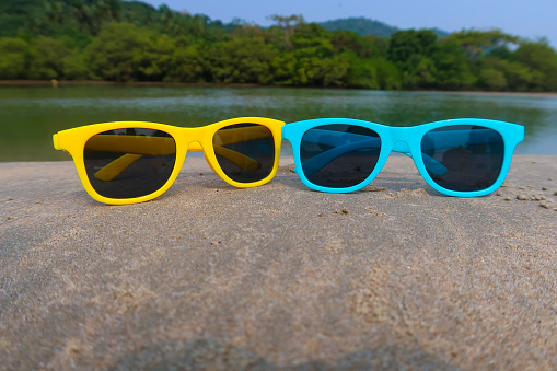 Stock photo showing two pairs of children's sunglasses, these brightly coloured blue and yellow glasses are placed next to each other on top of a rock at a beach in india, the water is reflecting the trees in the background, and the sky is blue on this sunny day.
