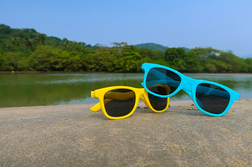 Stock photo showing two pairs of children's sunglasses, these brightly coloured blue and yellow glasses are stacked on top of each other on top of a rock at a beach in india, the water is reflecting the trees in the background, and the sky is blue on this sunny day.
