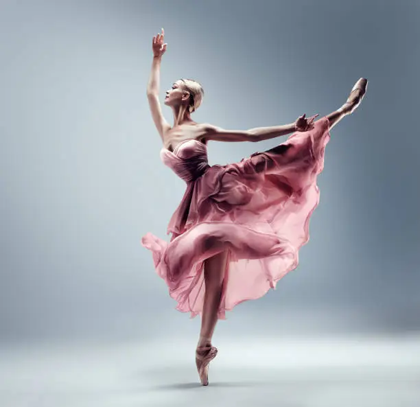 Ballerina in Pink Chiffon Dress jumping Split. Ballet Dancer in Silk Gown Pointe Shoes. Graceful Woman in Tutu Skirt dancing over Gray Studio Background