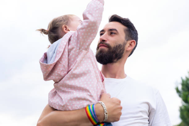 Father with a rainbow bracelet holding a little girl outdoors Father with a rainbow lgbt bracelet holding a little girl outdoors gay daddy stock pictures, royalty-free photos & images