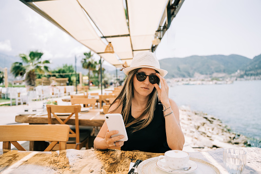 Young girl female lady in sunglasses looking at her mobile smart phone gadget in a street seaside cafe restaurant coffee shop with scenery mountains in the background. Hello summer holiday vacation.