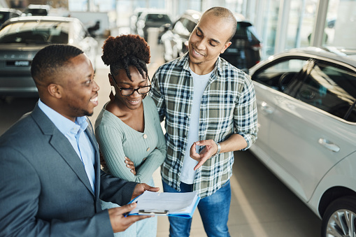 Happy African American couple  going through plans with car salesperson while buying a new car in a showroom.