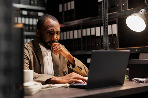 Worried detective analyzing crime case on laptop and thinking about evidence. Concerned african american policeman sitting at workplace desk in office full of racks with folders and files