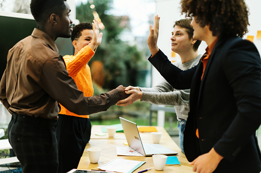 Young entrepreneurs congratulate each other, shake hands, and give a high-five after studying the planning of their startup