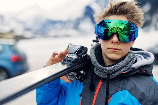 Teenage boy aged 13 is carrying skis on winter day. The boy is wearing modern mirrored ski goggles.\nCanon R5
