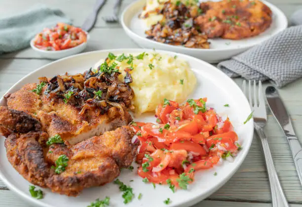 Delicious homemade dinner or lunch with breaded cutlet or pork chop. Served with mashed potatoes, roasted onions and tomato salad on plate with cutlery. closeup and front view