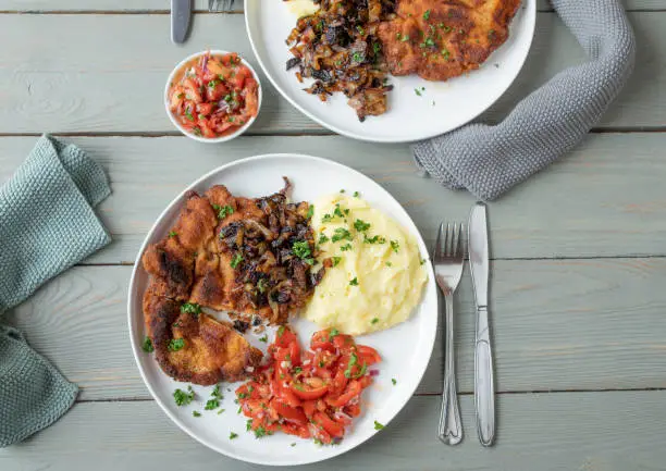 Delicious homemade dinner or lunch. Traditional cuisine with a pan fried and bread cutlet. Served with roasted onions, mashed potatoes and tomato salad on plates with cutlery and napkins on wooden table background. Flat lay