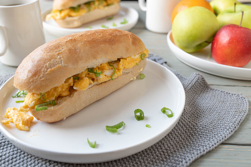Homemade breakfast sandwich with french baguette bun, scrambled eggs and chives. Served ready to eat on a kitchen table in the morning with a cup of coffee.