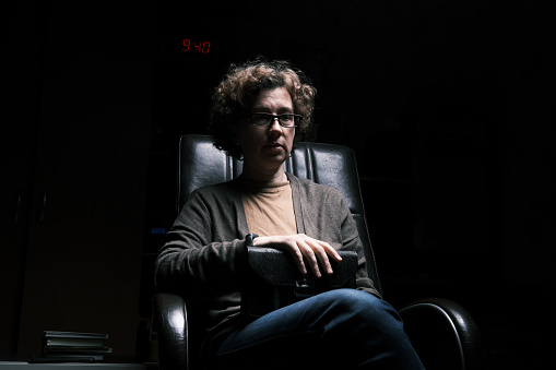 Sad mature woman sitting in the armchair in the dark room and holding a purse in her hands