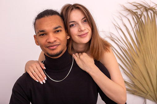 Portrait of happy beautiful multiracial couple, in black clothes, are sitting near the wall in the room, smiling, look at camera. Close up. Copy space