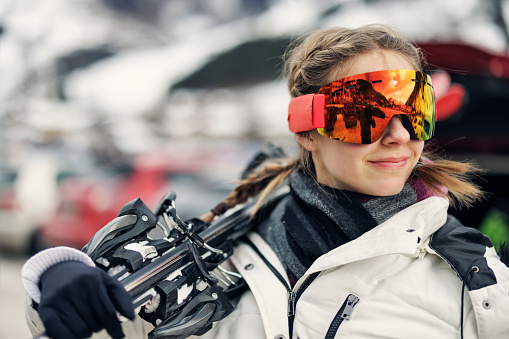 Teenage girl aged 17 is carrying skis on winter day. The girl is wearing modern mirrored ski goggles.\nCanon R5