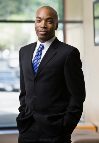 Portrait of a businessman of African descent standing in a lobby with his hands in his pockets.  He is wearing a suit and looking at the camera with a serious expression.  Vertical shot.