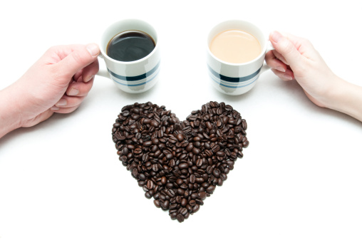 Two hands holding coffee cups, with a heart-shaped pile of coffee beans in the middle.