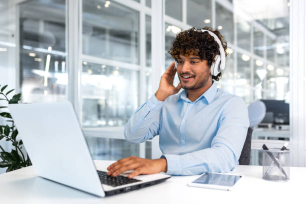 istock Young successful programmer inside modern office working with laptop, man in blue shirt smiling and happy, listening online music in headphones, audio books and podcasts, businessman satisfied work 1473165615