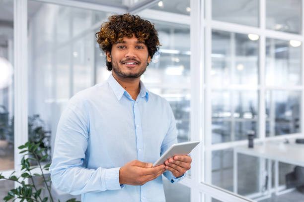 istock Portrait of young programmer with tablet computer inside office, hispanic man with curly hair smiling and looking at camera, man at workplace testing online applications standing near window 1473165693