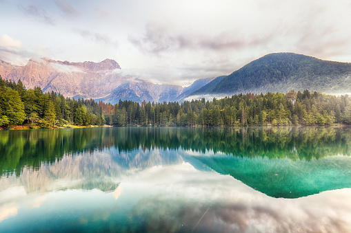 Scenic landscape with turquoise mountain lake in autumn valley against large snow mountains in low clouds in morning sunlight. Alpine lake with view to sunlit high snowy mountain range in low clouds.