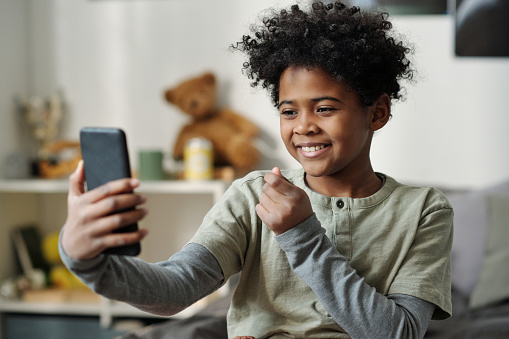 Happy schoolboy holding smartphone in front of himself and taking selfie or communicating in video chat while spending time at home