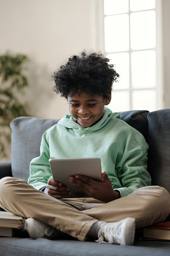 Smiling schoolchild with tablet watching online video or movie while sitting on couch in living room and preparing homework