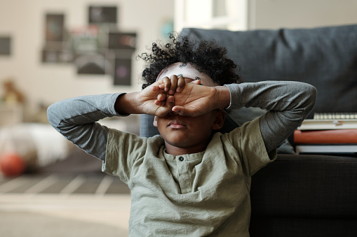 Offended or tired African American boy in grey pajamas hiding his face with hands while sitting by couch on the floor of living room