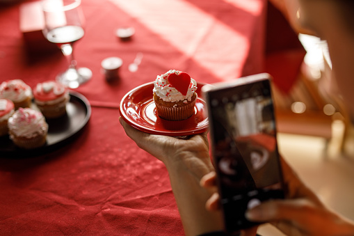 Close up shot of unrecognizable young woman sitting at table and using smart phone when taking photos of a delicious Valentine's day themed cupcake.