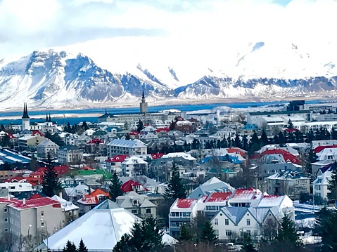 The town of Reykjavik with snow on the rooftops of the homes. There is a view of snow cap mountains in the distance.