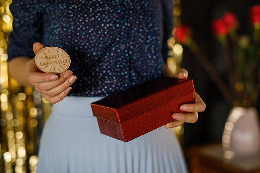 Midsection of unrecognizable young woman holding a gift box and wooden ornament engraved with words ''Happy anniversary''.