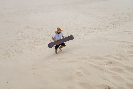 Man with a sand board wearing a straw hat walking on a sand dune in Balneario Atlantico beach, Arroio do Sal, RS, Brazil