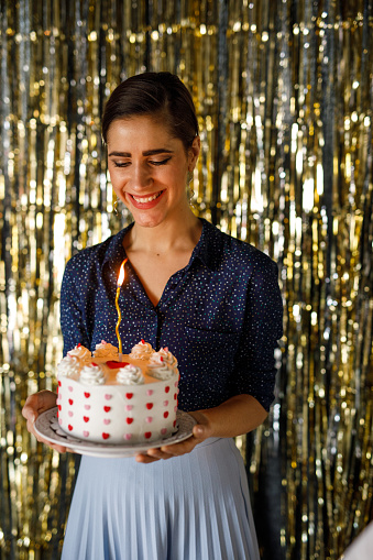 Portrait of joyful young woman standing against the gold colored tinsel curtain and making a wish before blowing out the birthday candle on the cake that she is holding.