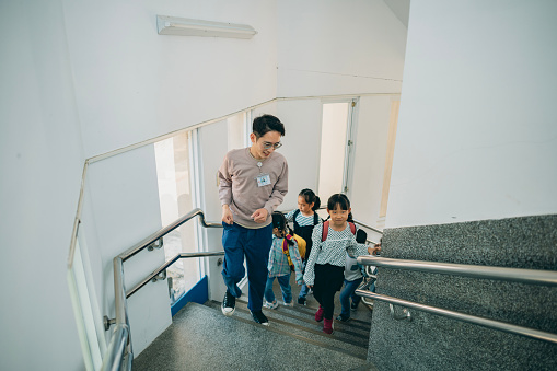 A preschool teacher in Asia is guiding children to line up in an orderly manner and climb the stairs to get ready to enter the classroom. During the process, the teacher reminds the children to stay safe and discusses the activities planned for the day.