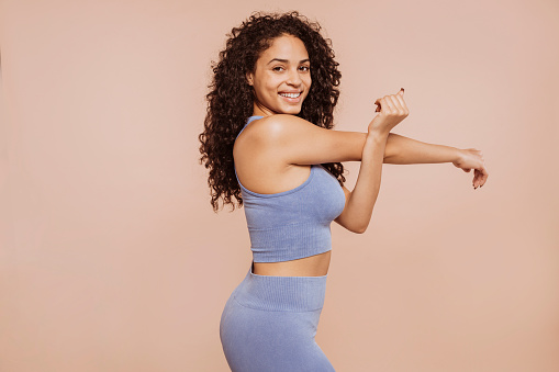 Side view of young cheerful multiracial curly-haired brunette sportswoman wears blue sportswear, stretching arm at studio isolated over beige background.