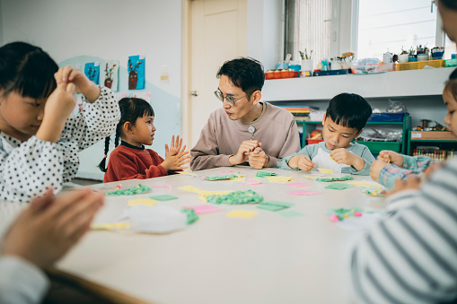 An Asian preschool teacher is guiding the children in a handicraft activity, encouraging them to unleash their creativity to complete an art piece of a tree, and extending the lesson to include activities about loving the earth and promoting sustainable development.