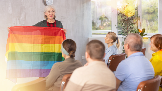 Smiling elderly female tutor participating in seniors education program, talking about international LGBT social movements to group in auditorium, showing colorful rainbow flag