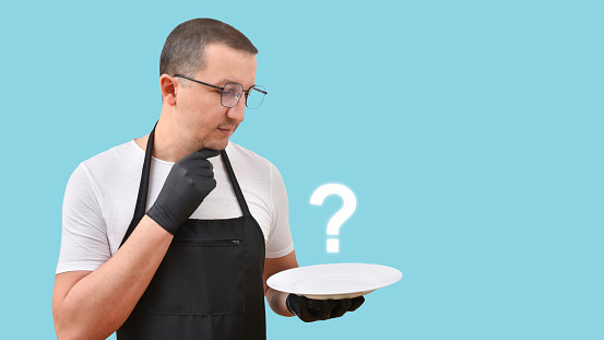 A cook in an apron with a plate in his hands thought about the dish on a blue background. The concept of cooking.