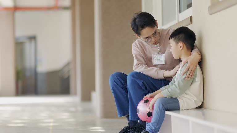 An Asian preschool teacher is sitting outside the classroom, having a conversation with a discouraged child. She is attentively listening and providing comfort to the child.