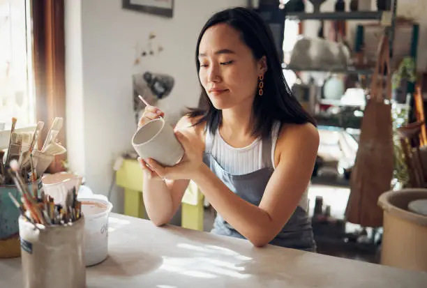 Photo of Pottery, art and sculpture with a japanese woman in a studio for design or a creative hobby as an artisan. Manufacturing, pattern or artist with a female potter sitting in her workshop  as a sculptor