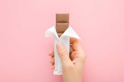 Young adult woman hand holding brown chocolate bar on light pink table background. Pastel color. Closeup. Sweet snack in opened white pack. Top down view.