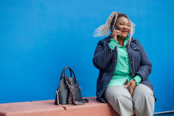 Calling Friends After Work A shot of a mid-adult female sitting outside against a blue wall with her handbag beside her. She is smiling whilst talking on the phone and wears smart-casual clothing. older woman purse stock pictures, royalty-free photos & images